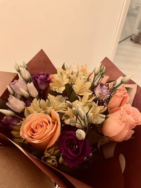 A floral bouquet featuring peach roses with delicate, tightly wound petals, complemented by deep purple lisianthus with ruffled petals, and pale cream alstroemeria with slender, open blooms. The arran
