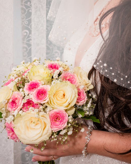 A close-up of a bride's hands holding a floral bouquet arrangement consisting of large, creamy white roses and smaller pink roses with delicate white baby's breath accents. The bouquet is wrapped in e