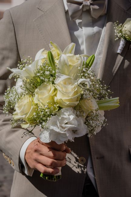 A close-up view of a groom holding a bridal bouquet composed of creamy white roses, white lilies, and delicate baby's breath, with hints of green foliage. The bouquet is wrapped in white ribbon, and t