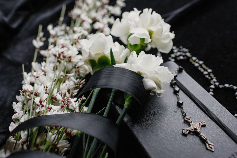 A close-up of a floral arrangement featuring white carnations and small white blossoms, accompanied by green stems, all resting on a bed of black wrapping material. The flowers are fresh with delicate