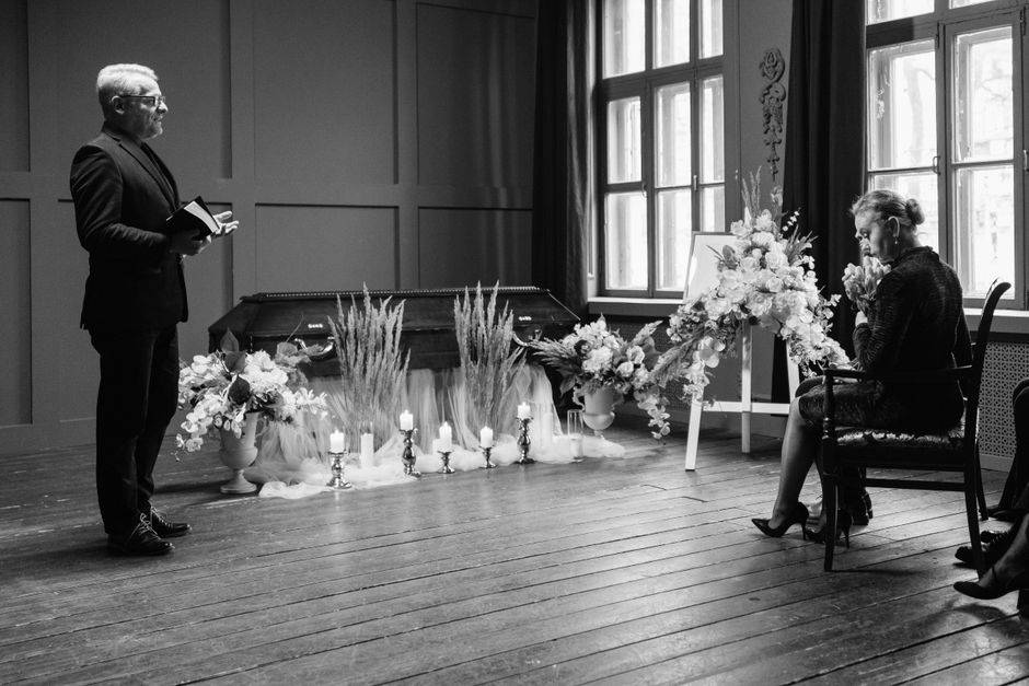 A black-and-white photograph capturing a funeral scene with floral arrangements. On the left, a man dressed in a dark suit holds a small book or program, standing quietly. To the right, a woman seated