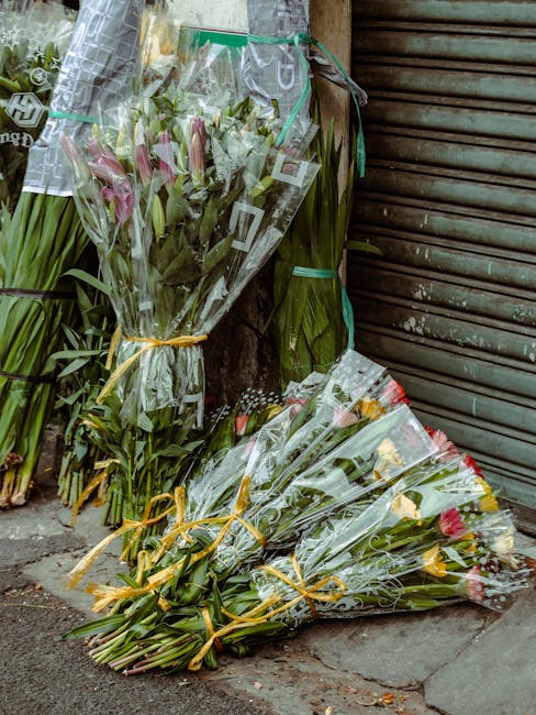 Several bouquets of fresh flowers, including pink lilies, yellow roses, and mixed floral arrangements, wrapped in clear cellophane with ribbon ties, are displayed against a weathered brick wall and a