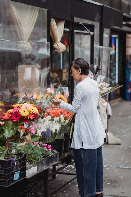 A woman with short black hair, dressed in a white long-sleeve top and dark blue jeans, is standing outside a flower shop, selecting from a vibrant display of fresh flowers arranged in black buckets. T