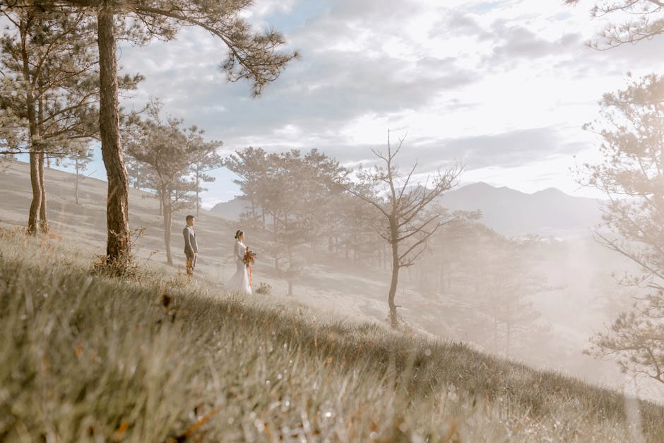 A bride holding a vibrant bouquet of red, orange, and yellow flowers, dressed in a white wedding gown, standing on a grassy hillside amidst tall pine trees with distant mountains in the background. Th
