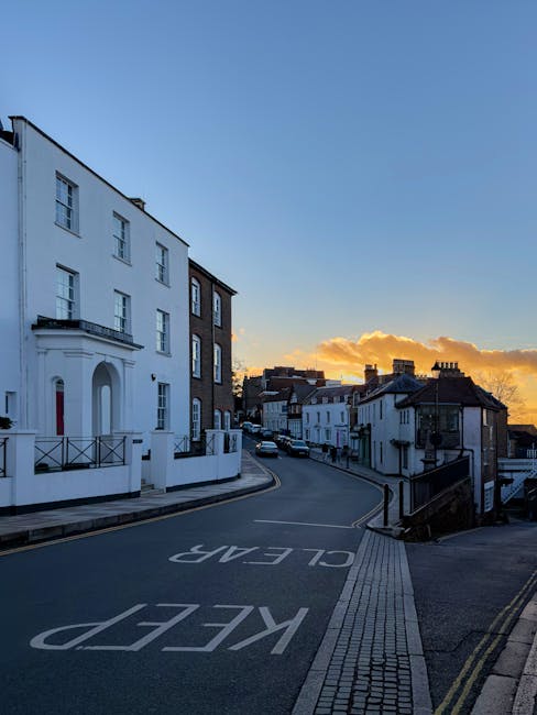 This image features a gentle hillside street in Harrow on the Hill at sunset, with a row of elegant white and dark brick buildings lining the road. The sky is a gradient of soft blue transitioning int