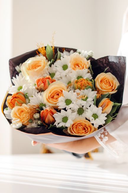 A hand holding a bouquet of fresh flowers, featuring large peach-colored roses and white daisies with green centers, arranged with dark purple wrapping paper and a white ribbon, set against a neutral 