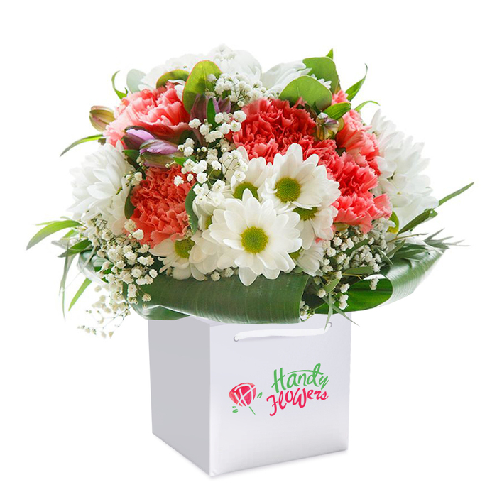 Presented upright in a crisp, minimalist white gift bag, this vibrant bouquet embodies a celebration of texture, colour, and craftsmanship typical of North Harrow's distinguished florists. The arrangement's focal point is the coral and soft peach carnations-recognizable by their abundant, frilled petals which create visual richness and tactile allure. Interspersed between them, bold white daisies with radiant yellow-green centers enliven the arrangement with brightness, echoing the spirit of nearby North Harrow markets on a lively morning. Wispy baby's breath blossoms are woven throughout, providing a gentle, cloud-like softness that balances the more robust blooms. On the bouquet's left, several small purple-tipped buds lend an added layer of intrigue and evolving beauty. Surrounding this floral medley, broad, glossy deep green leaves fan out in an elegant collar, underpinning the bouquet with structure and a lush natural feel. The handcrafted, rounded shape speaks to the experienced hand of a North Harrow florist, while the thick white ribbon handles of the bag hint at both practicality and refined presentation. Whether for a birthday surprise on Pinner Road, a congratulatory gesture, or a heartfelt thank you, this arrangement encapsulates the promise of freshness, quality, and the personal touch of a shop truly rooted in the local area.