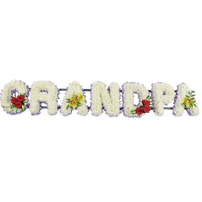 A large funeral flower display spells the word "GRANDPA" in striking capital letters, laid out in a straight line against a plain white background so that the details of the floristry can be clearly imagined. Each letter is built on a hidden frame and completely carpeted in small, pristine white chrysanthemum heads, tightly grouped so there are no gaps, creating a smooth, padded surface that suggests softness and care. The petals catch the light gently, giving the tribute a quiet glow, like the calm of an overcast day in North Harrow's memorial gardens. Along the very edge of each letter, a slim lavender-purple ribbon forms a neat outline, adding definition and a hint of colour without disturbing the overall white purity. The florist has introduced bolder accents at key points: the curve of the initial "G" houses a rounded cluster of deep red roses, nestled among short stems of dark green foliage; a matching cluster appears within the bowl of the "D," balancing the design and framing the central part of the word with love. On the first and last "A," elegant sprays of yellow-green orchids, some with a touch of reddish-brown in their throats, arch gently over the white base, lending a refined, almost regal touch. Narrow connecting bars support the letters from behind, ensuring the word remains perfectly aligned when displayed on a stand during a funeral, wake, or graveside service in North Harrow, Harrow Weald, or the surrounding area. The finished piece feels both formal and deeply personal, combining the peace of white chrysanthemums with the passion of roses and the quiet sophistication of orchids to honour a grandfather's life and legacy.